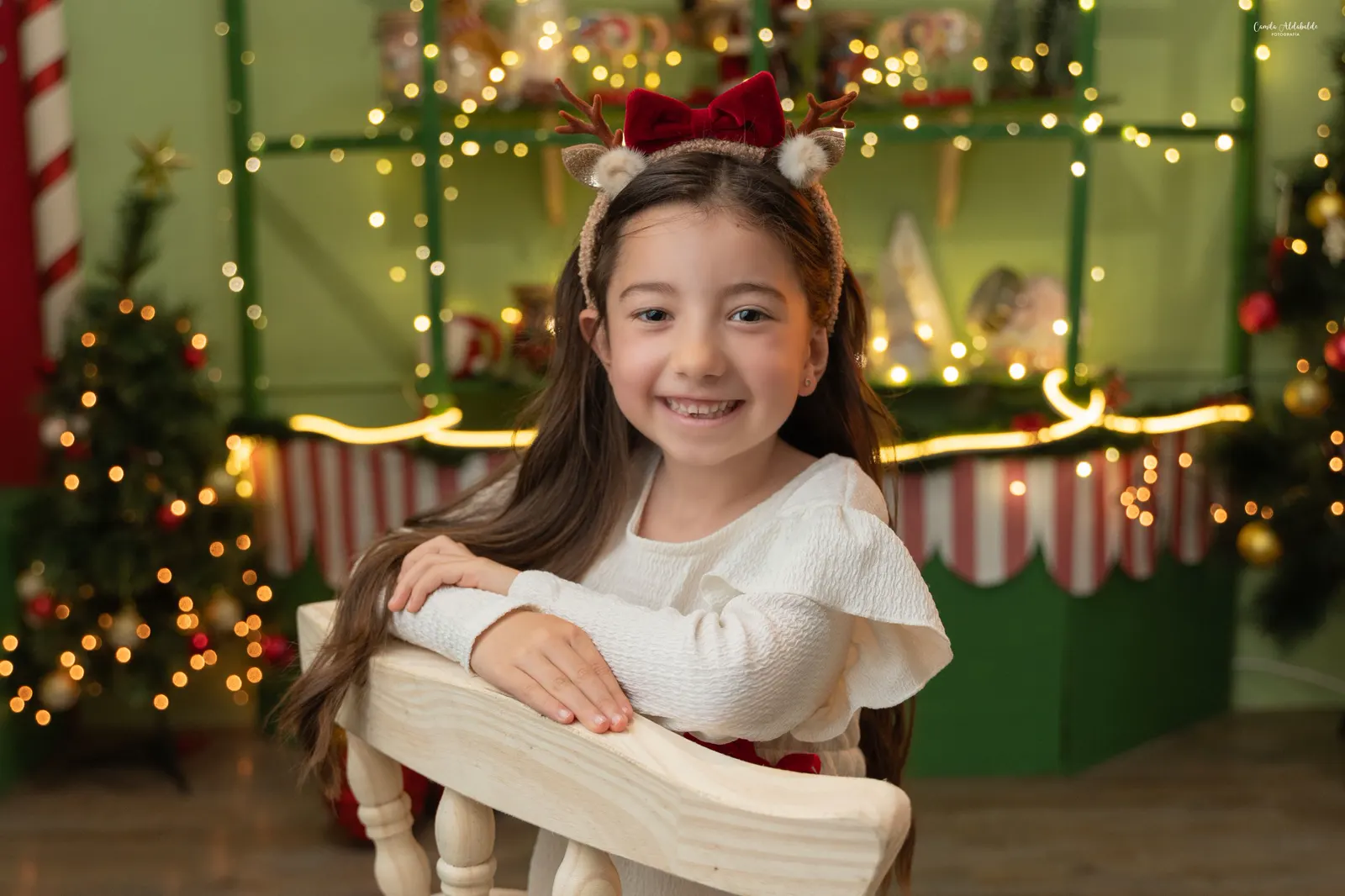 Niña sonriendo apoyada en una silla de madera, con vincha de reno, en el set navideño de tienda de dulces